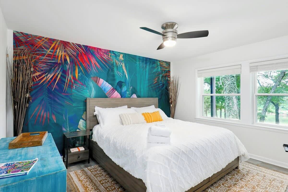 A bedroom with a bed, a ceiling fan, a window, and a large abstract painting on the wall.