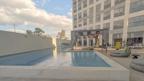 A swimming pool with lounge chairs and a covered area, surrounded by a modern building. - Depa de lujo en Dúo 24 | Alberca, gym y seguridad (Zapopan)