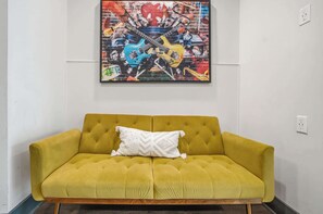 Ein senfgelbes Sofa mit einem weißen Zierkissen vor einer weißen Wand, darüber ein farbenfrohes abstraktes Gemälde.