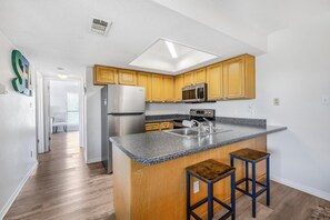 A modern kitchen with wooden cabinets, a granite countertop, a stainless steel refrigerator, and a microwave. - 🏖️ Romar Village 621 | 2BR Orange Beach Condo | Near Beach + Sleeps 4 (Orange Beach)