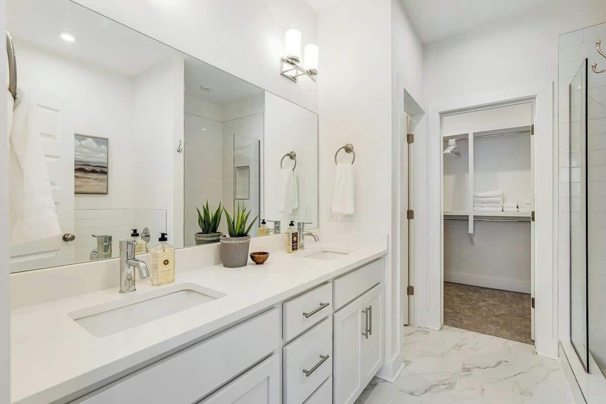 A modern bathroom with a large mirror, two sinks, a shower, and a walk-in closet.