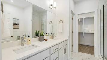 A modern bathroom with a large mirror, two sinks, a shower, and a walk-in closet.