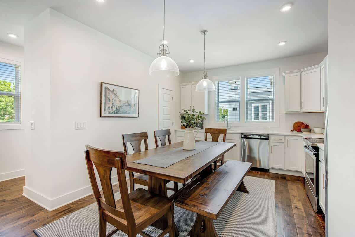 A dining area with a wooden table and chairs, a kitchen with white cabinets, and pendant lighting.