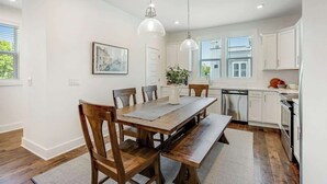 A dining area with a wooden table and chairs, a kitchen with white cabinets, and pendant lighting.