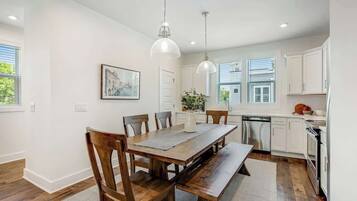 A dining area with a wooden table and chairs, a kitchen with white cabinets, and pendant lighting.