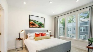 A modern bedroom with a bed, bedside lamps, a painting, and large windows.