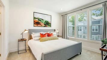 A modern bedroom with a bed, bedside lamps, a painting, and large windows.
