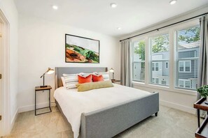 Un dormitorio moderno con cama, lámparas de noche, un cuadro y ventanas grandes.