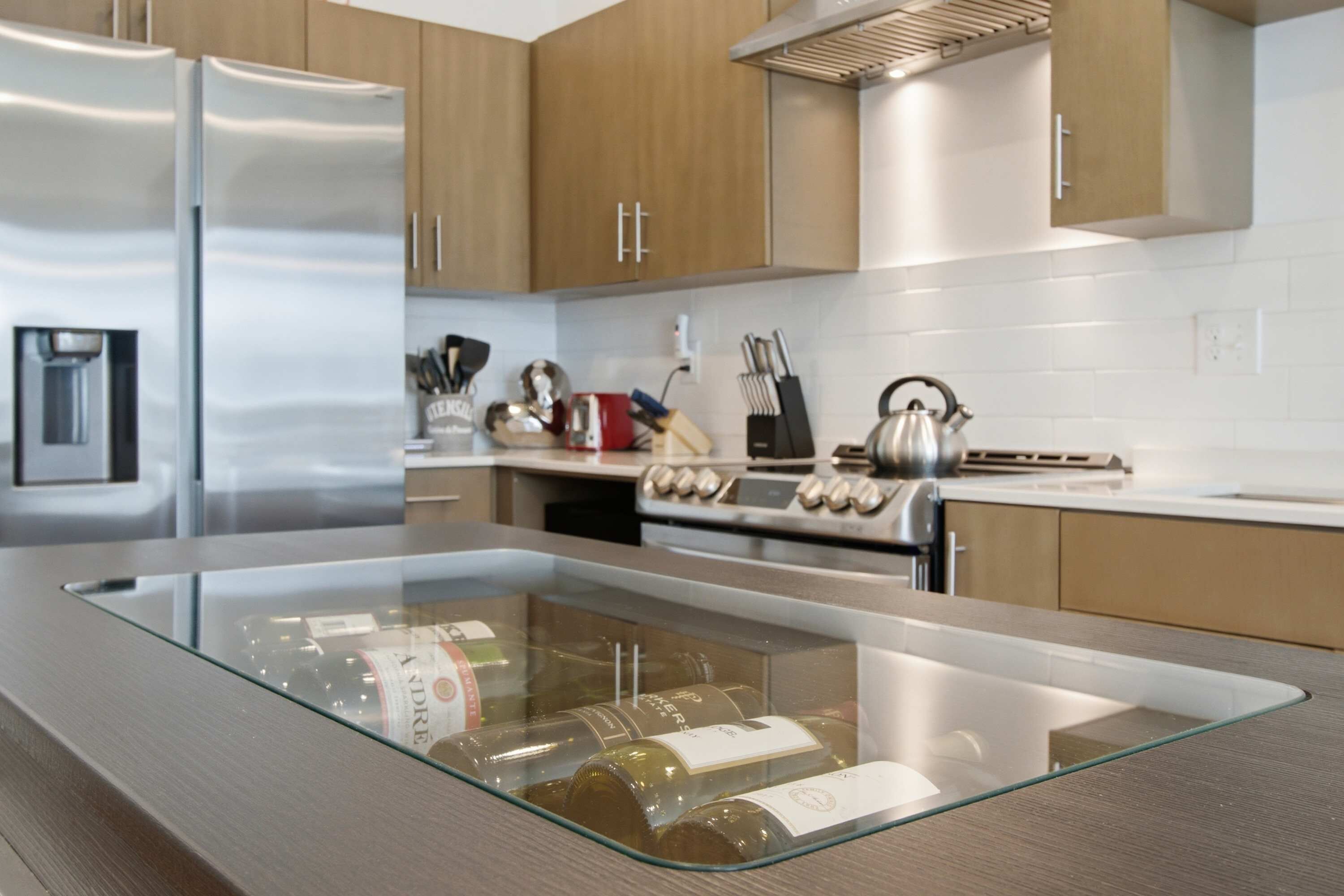 Une cuisine moderne dotée d’appareils électroménagers en acier inoxydable, de meubles en bois et d’un îlot central avec un plateau en verre, sur lequel sont disposées des bouteilles de vin.