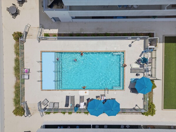 An aerial view of a rectangular swimming pool with a surrounding deck, lounge chairs, umbrellas, and a small garden area. - Chic Corner Condo Escape with Seasonal Pool Access (Nashville)