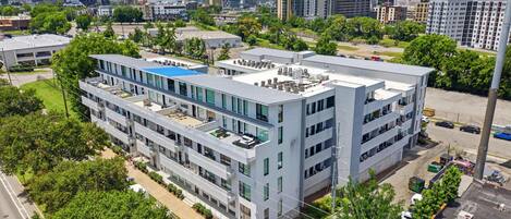 A modern multi-story building with a rooftop terrace, surrounded by greenery and a parking area.