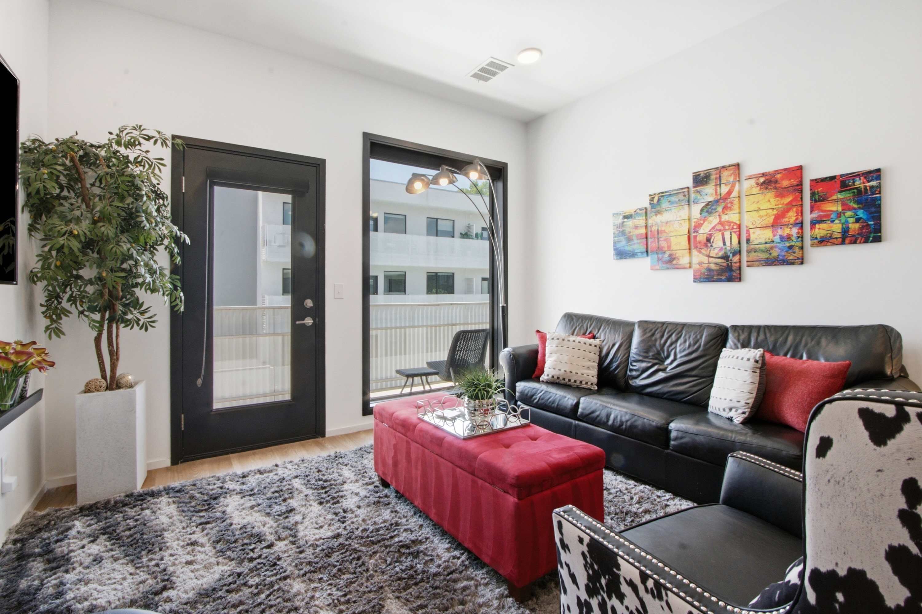 A modern living room with a black leather sofa, a red ottoman, and a black and white patterned chair. There are three abstract paintings on the wall, a potted plant, and a large window with a view of a building.