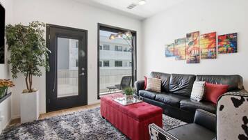 A modern living room with a black leather sofa, a red ottoman, and a black and white patterned chair. There are three abstract paintings on the wall, a potted plant, and a large window with a view of a building.