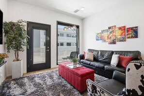 Een moderne woonkamer met een zwarte leren bank, een rode poef en een fauteuil met een zwart-wit patroon. Op de muur hangen drie abstracte schilderijen, er staat een potplant en is een groot raam te zien met uitzicht op een gebouw.