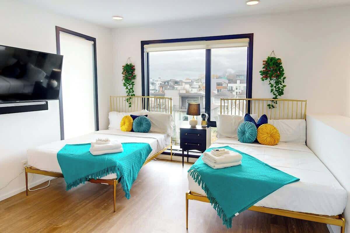 A modern hotel room with two beds, a TV, a large window, and a view of the city.