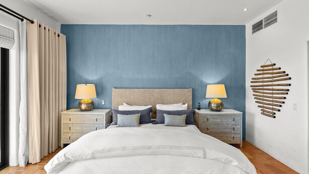 A bedroom with a bed, two bedside tables, a headboard, a wall-mounted light fixture, and a window with curtains. - Newly Renovated Condo With Ocean View Luxury Amenities in Beachfront Resort Walk to Downtown (Cabo San Lucas)