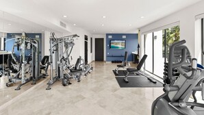 Ein modernes Fitnessstudio mit verschiedenen Trainingsgeräten, einem großen Fernsehbildschirm und Blick auf Palmen durch die Fenster.