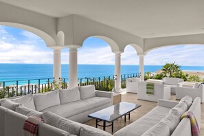 A covered outdoor seating area with white sofas and a table, overlooking a beach and the ocean. - Breathtaking Villa Boasting a Private Putt Chip Course Gym Tennis Court Resort Access More (San José del Cabo)