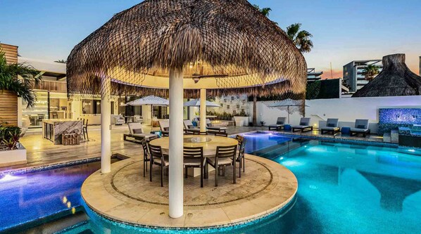 A pool area with a round table, lounge chairs, and a thatched umbrella. - Ultra Exclusive Beachfront Villa Just Steps From the Sand Butler Included (San José del Cabo)
