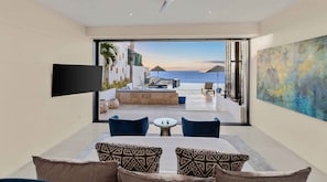 A modern hotel room with a large window, a flat-screen TV, a dining area with a table and chairs, and a view of the sea. - Ultra Exclusive Beachfront Villa Just Steps From the Sand Butler Included (San José del Cabo)