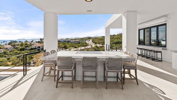 A modern outdoor dining area with a panoramic view, featuring a large table and chairs, and a glass railing. - Brand New Villa BIG Ocean View Gym Close to the Beach (San José del Cabo)