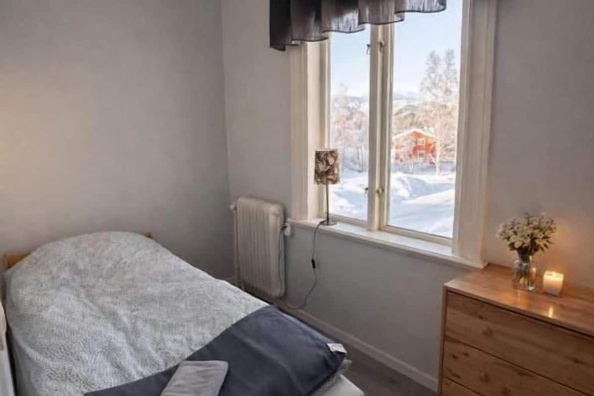 Un dormitorio con cama, ventana con cortinas, radiador, cómoda de madera y un florero con flores.