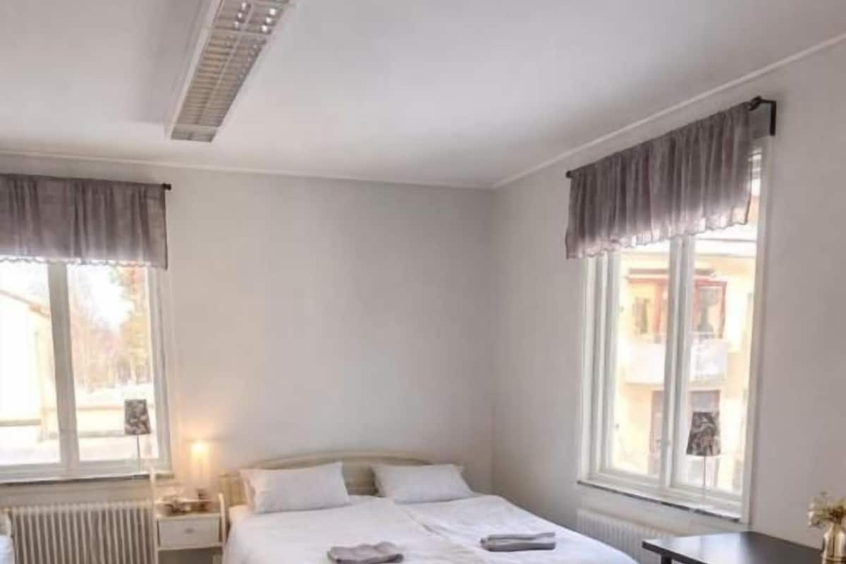 Un dormitorio con cama, mesitas de noche, escritorio, ventana con cortinas y radiador.