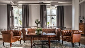 A room with a brown leather sofa, two armchairs, a round coffee table, and a large window with curtains. - Wärdshuset Liden (Liden)