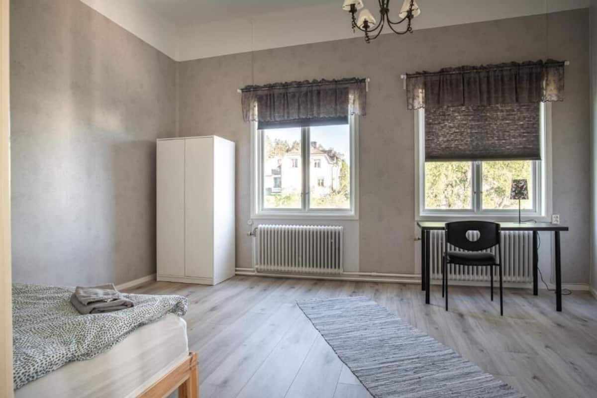 Un dormitorio con cama, radiador, escritorio con silla y dos ventanas con cortinas.