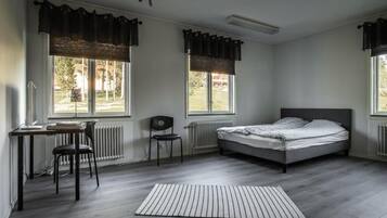 A bedroom with a bed, a desk, a chair, and two windows with curtains.