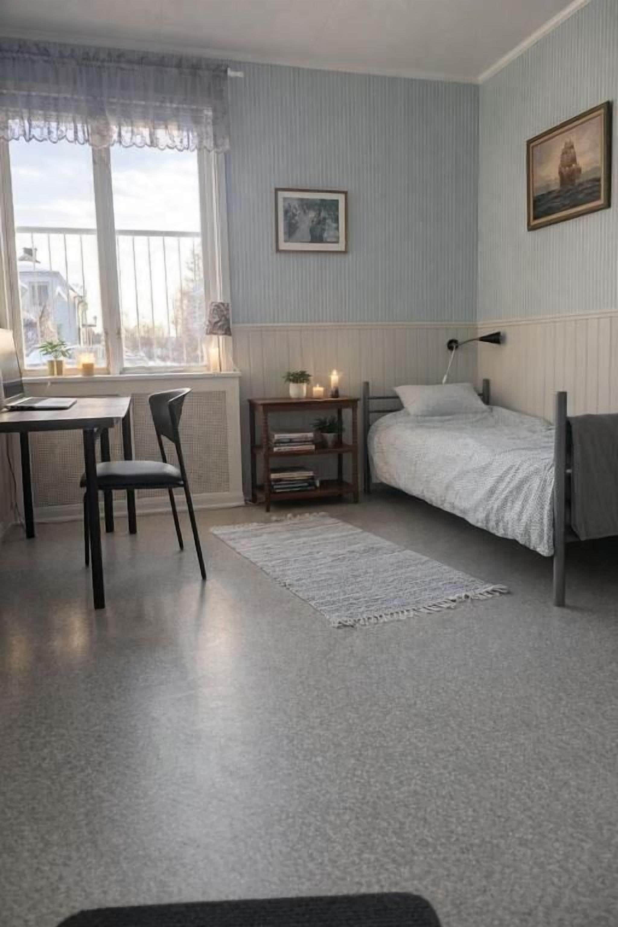 Un dormitorio con cama, escritorio, silla, mesita de noche con lámpara y ventana con cortinas.