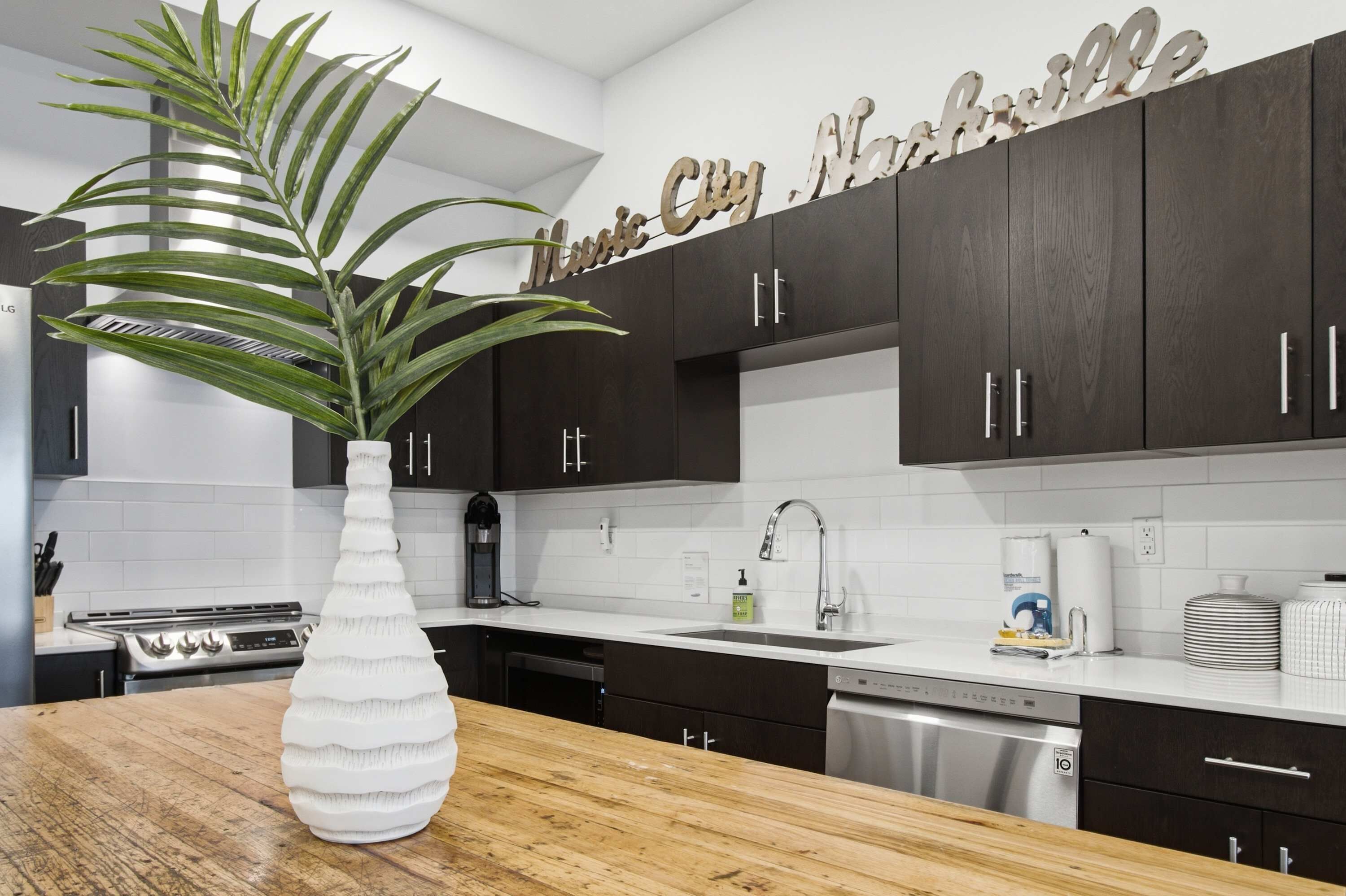 A modern kitchen with dark cabinets, a white countertop, a large white vase with a plant, and a sign above the cabinets.