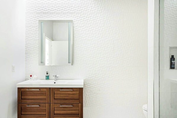 A bathroom with a white textured wall, a white sink with a mirror above it, and a glass shower enclosure. - Charming Condo Stay with Tasteful Decor, Sleeps 4 (Nashville)