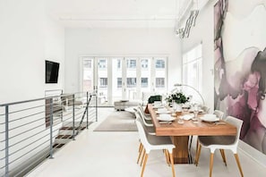 A modern dining area with a wooden table set for six, white chairs, and a large window with a view of buildings. - Charming condo with central AC, full kitchen, & tasteful decor (Nashville)