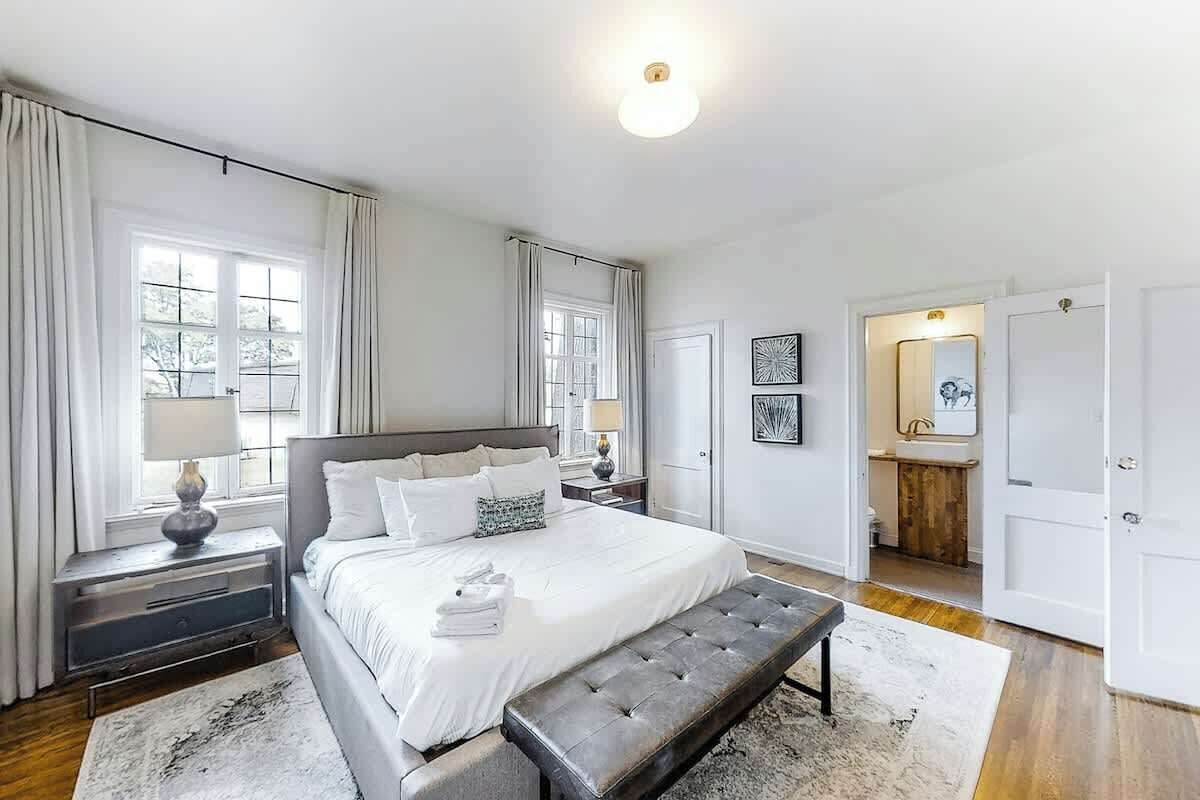 Ein Schlafzimmer mit einem Bett, Nachttischen, einer Bank, einem Fenster mit Vorhängen und einem durch eine offene Tür einsehbaren Badezimmer.