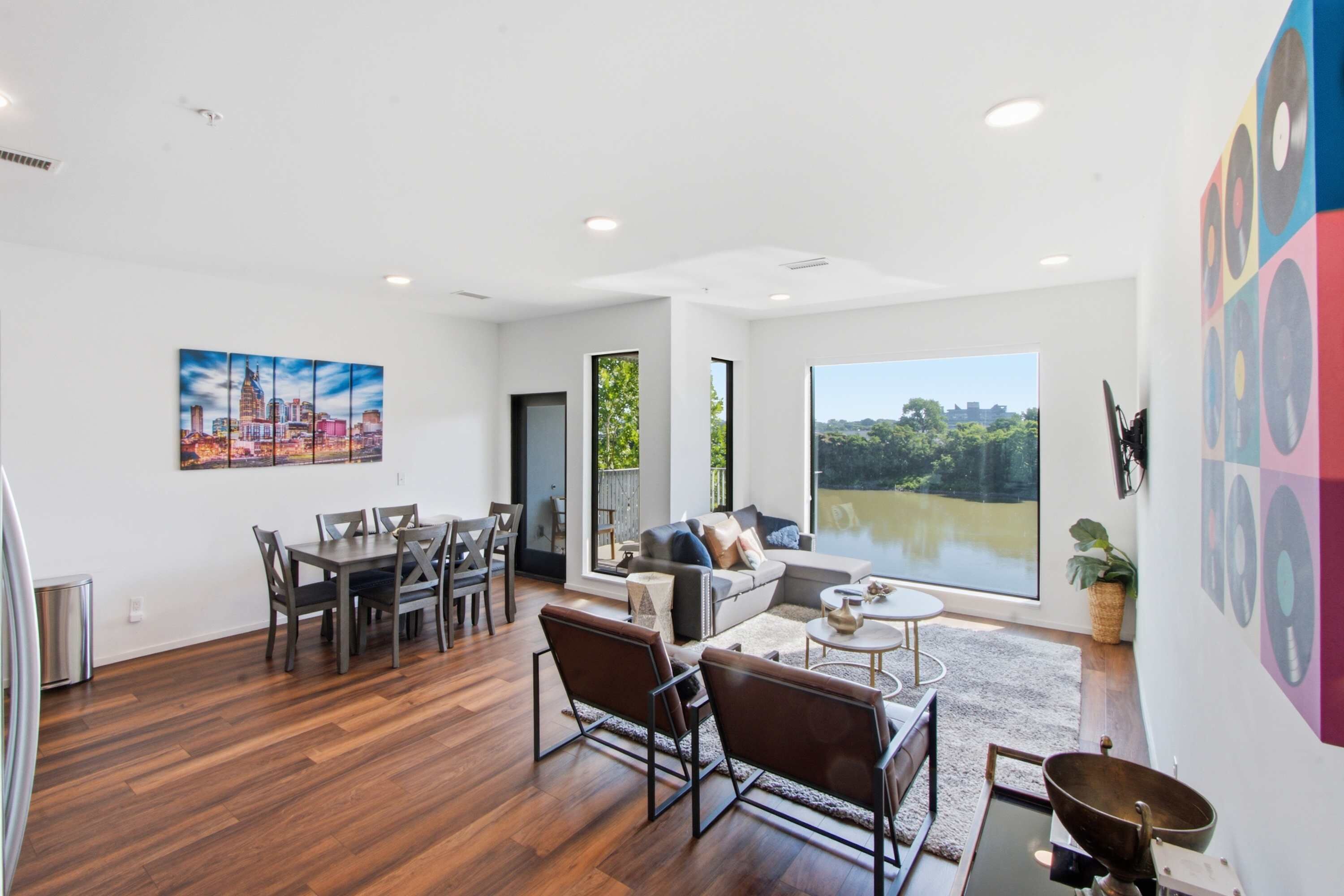 A spacious living area with a dining table, sofas, and a large window offering a view of greenery.