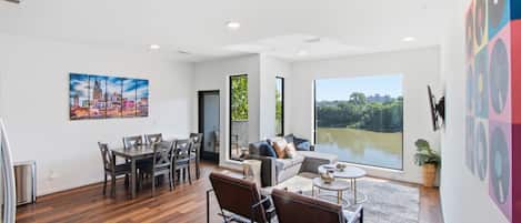 A spacious living area with a dining table, sofas, and a large window offering a view of greenery.
