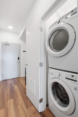 A laundry room with two stacked front-loading washing machines, a door with a handle, and a wooden floor. - Riverfront Condo Stay with Balcony, Near Downtown (Nashville)
