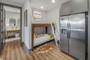 Ein kompaktes Hotelzimmer mit Etagenbett, einem modernen Kühlschrank, Holzboden und einem Badezimmer mit Spiegel und Waschbecken.