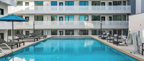 A modern hotel with a swimming pool, sun loungers, and a covered area with umbrellas.