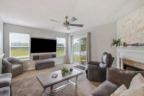 Een woonkamer met een flatscreen televisie, een plafondventilator, een open haard en een groot raam.