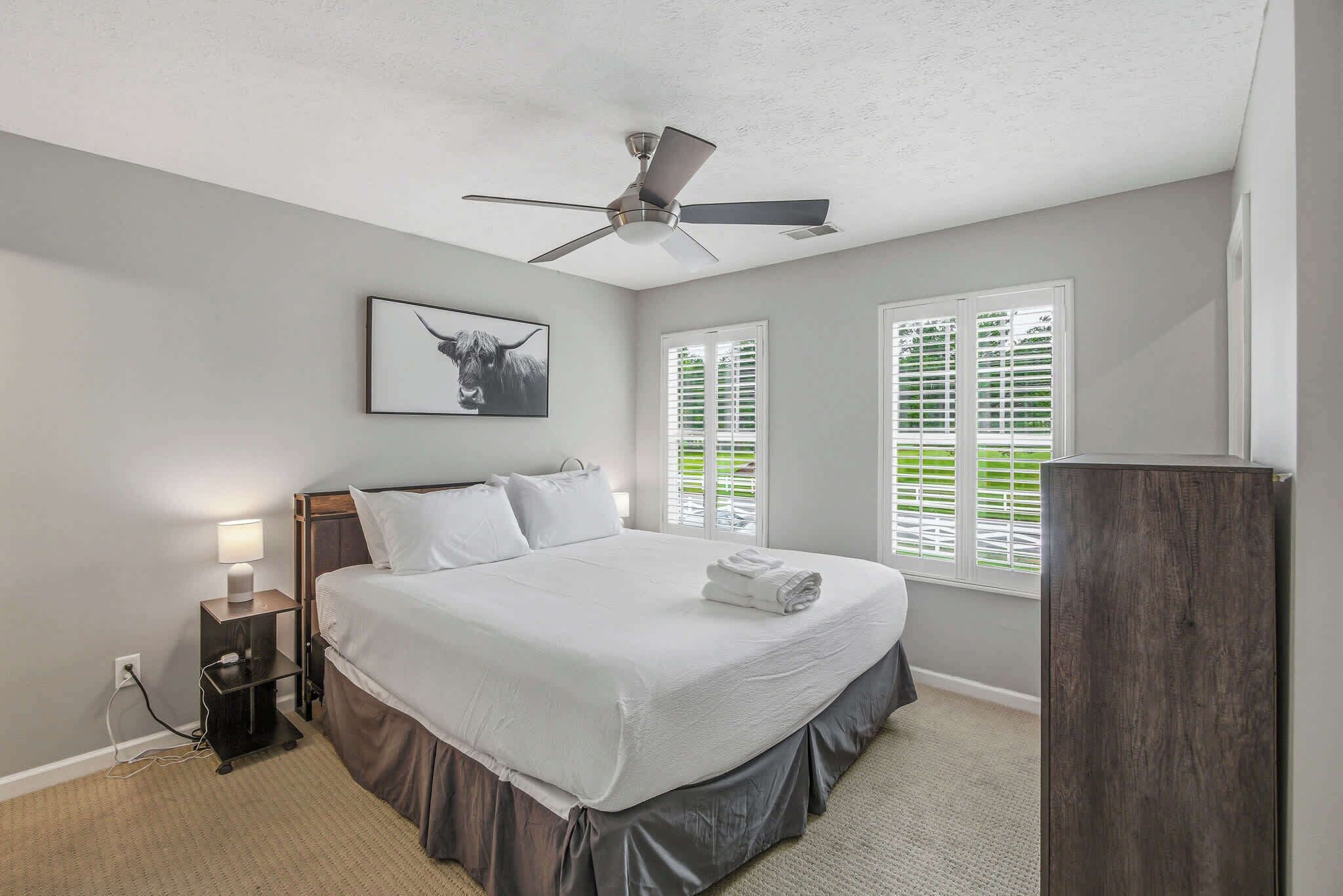 A bedroom with a bed, bedside table, lamp, ceiling fan, and windows with blinds.