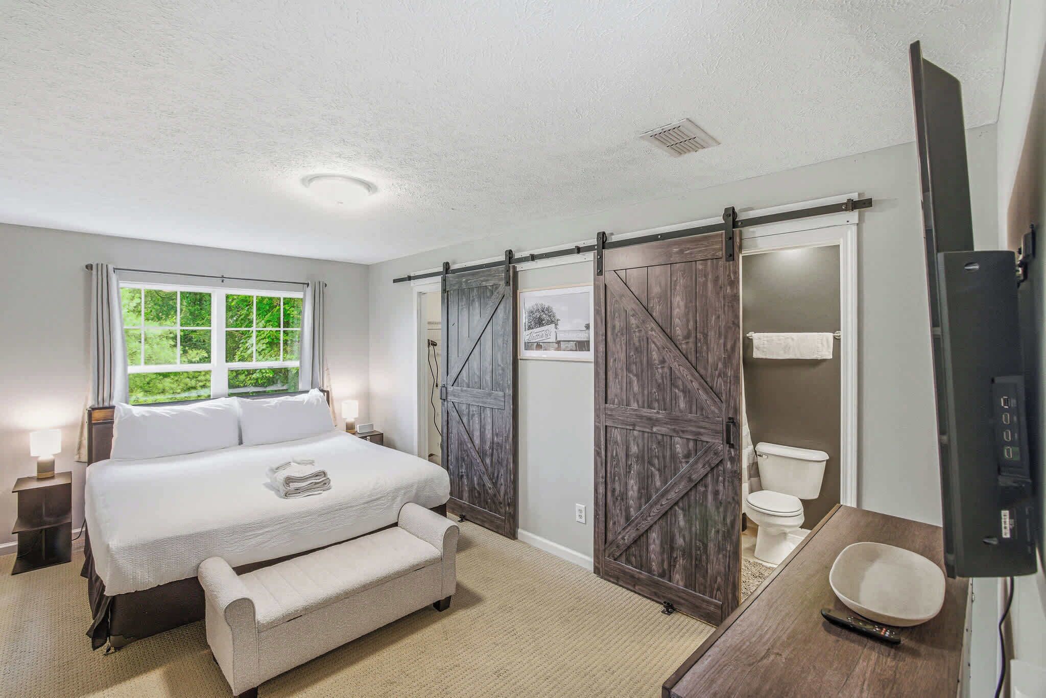 A bedroom with a bed, a bench, a window, a door, a toilet, and a television.