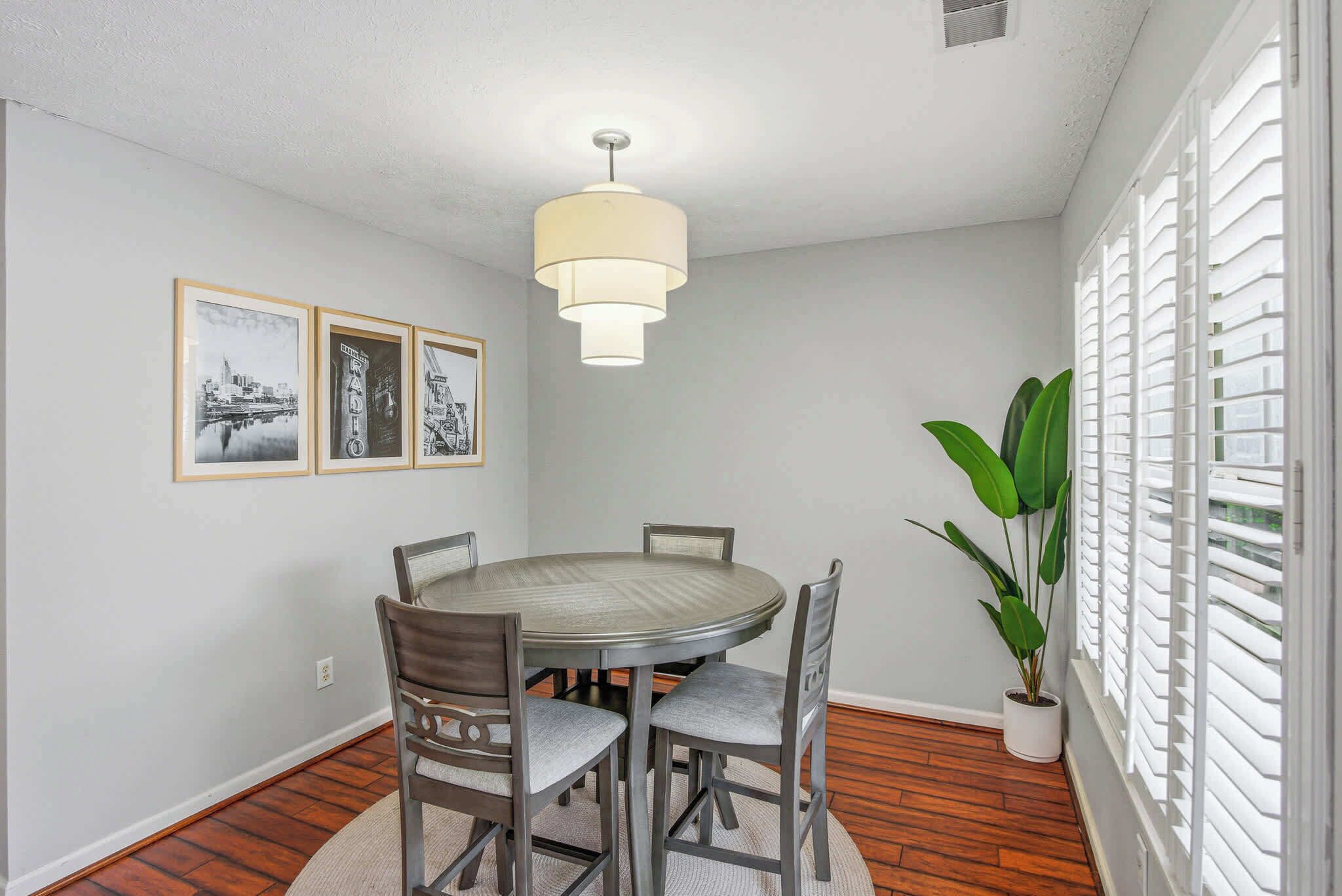 A dining room with a round table, four chairs, a hanging light fixture, and three framed pictures on the wall.