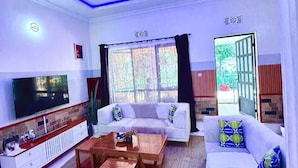 Una sala de estar con televisor, sofás, mesas auxiliares y un ventanal grande.