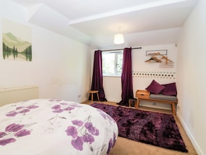 A bedroom with a bed, a window with curtains, a radiator, a hanging light fixture, and a wall-mounted shelf.
