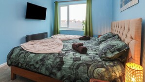 A bedroom with a bed, a radiator, a TV, a window with curtains, and a hanging light fixture. - Home by the sea (Llandudno)