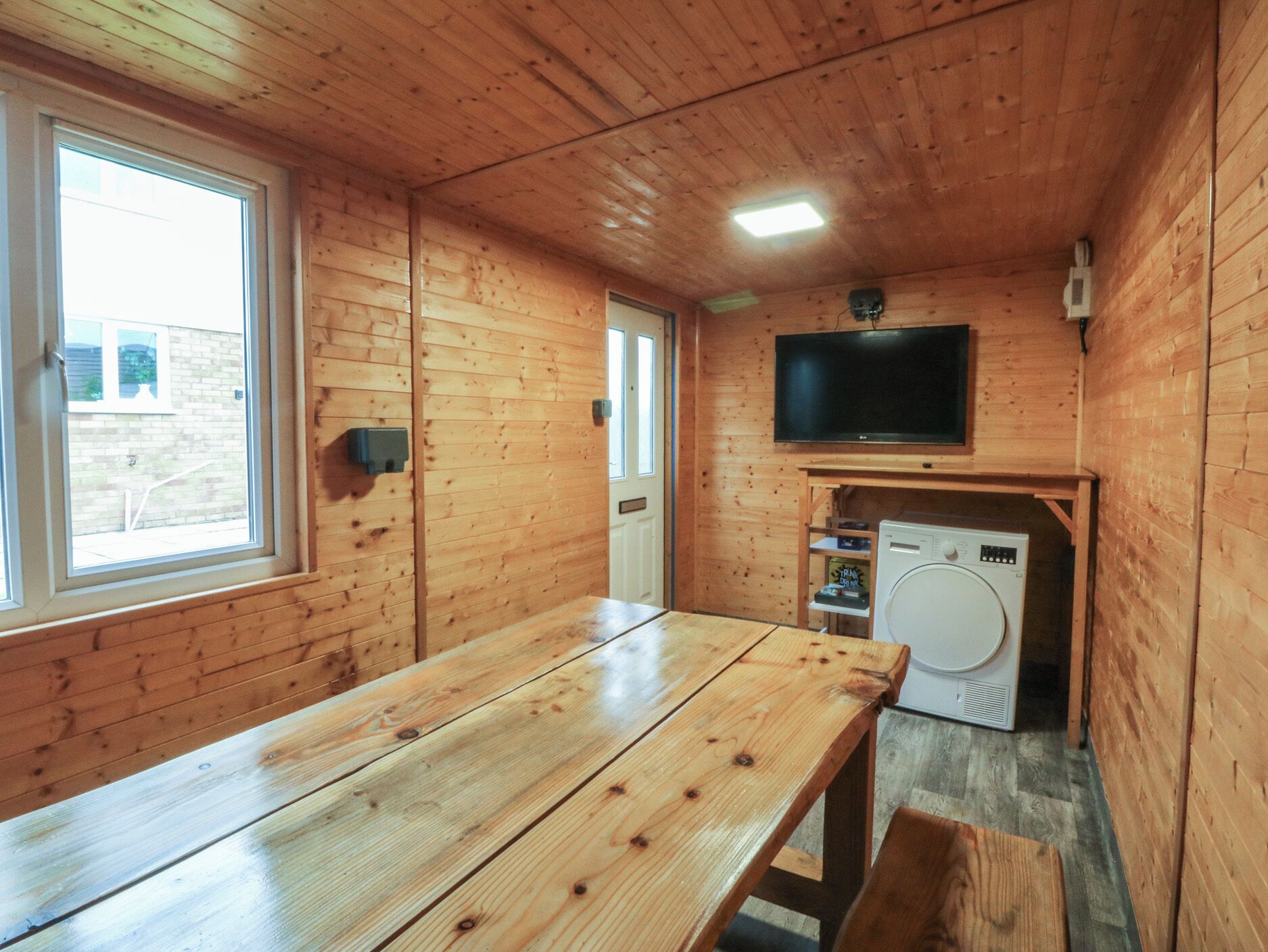 A wooden cabin interior with a wooden table, a washing machine, a television mounted on the wall, and a window.