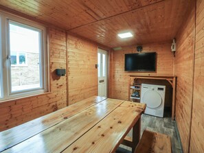A wooden cabin interior with a wooden table, a washing machine, a television mounted on the wall, and a window. - Home by the sea (Llandudno)