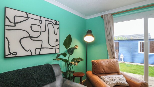 A living room with a grey sofa, a brown armchair, a floor lamp, a large abstract painting, and a view of a blue house through a glass door. - Home by the sea (Llandudno)